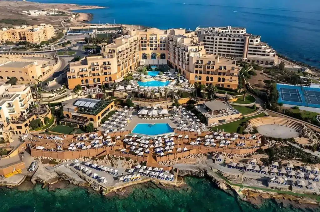 Corinthia Hotel St George's Bay exterior view with private sandy beach, turquoise Mediterranean waters, and luxury resort facade in St Julian's Malta